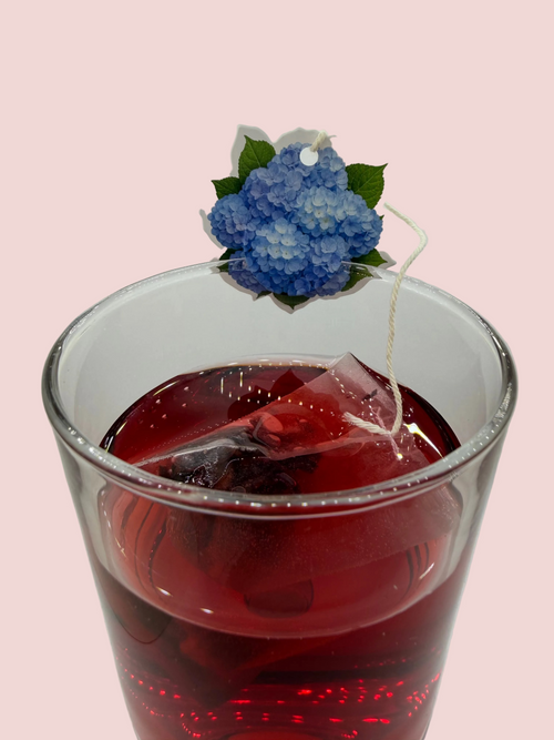 A glass cup filled with hibiscus tea and tea tag of blue hydrangea sitting on the rim of the cup.