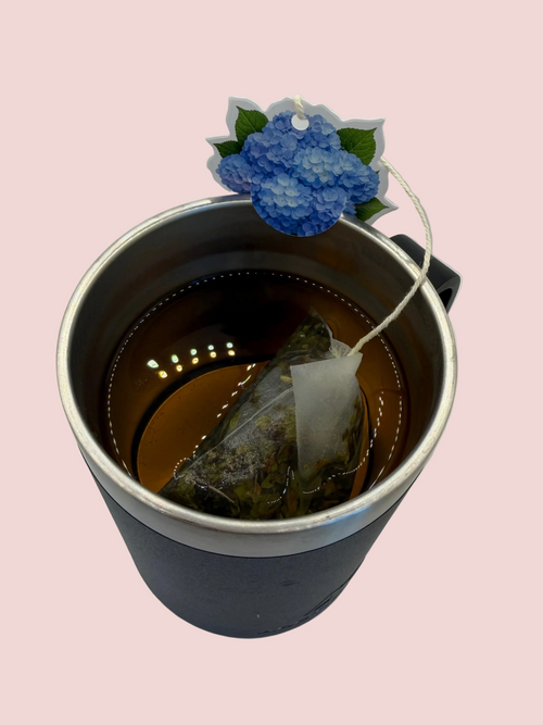 An insulated mug with a silver rim, filled with peppermint tea and tea tag of blue hydrangea sitting on the rim of the cup.