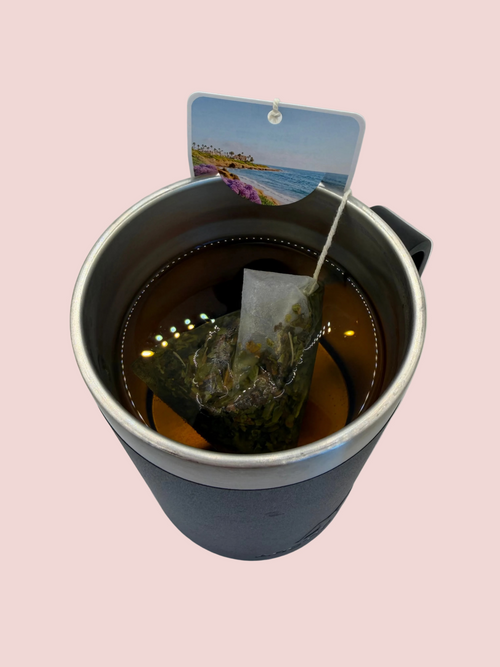 A travel mug filled with peppermint tea and a tea bag featuring a scenic view of La Jolla Cove sitting on the rim of the mug.