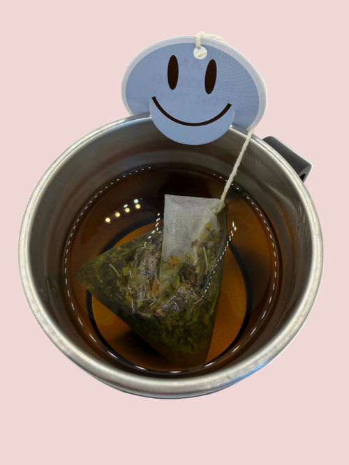 A stainless steel mug filled with peppermint tea with a blue smiley face tag sitting on the rim of the mug. 