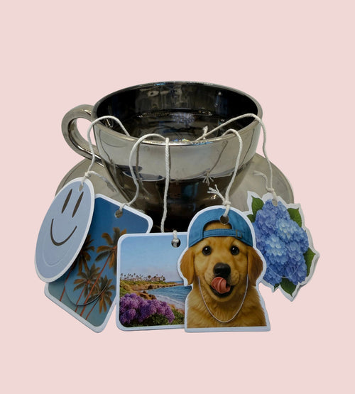 A silver teapot with a matching saucer, decorated with several colorful printed hanging tags featuring a smiley face, palm trees, La Jolla Cove, a golden retriever and blue hydrangea.